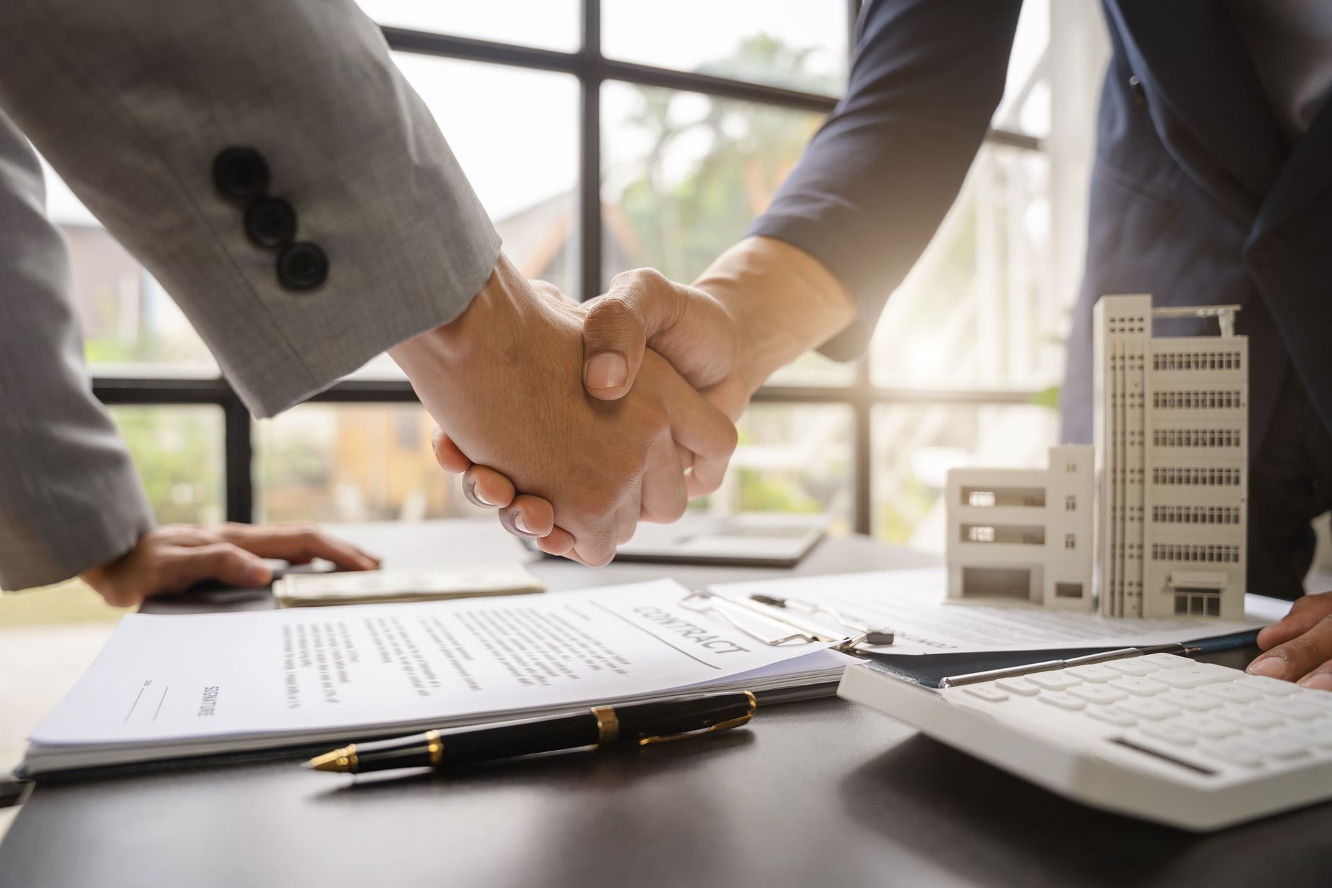 Business handshake over documents at a desk, with calculator and office building model in the background