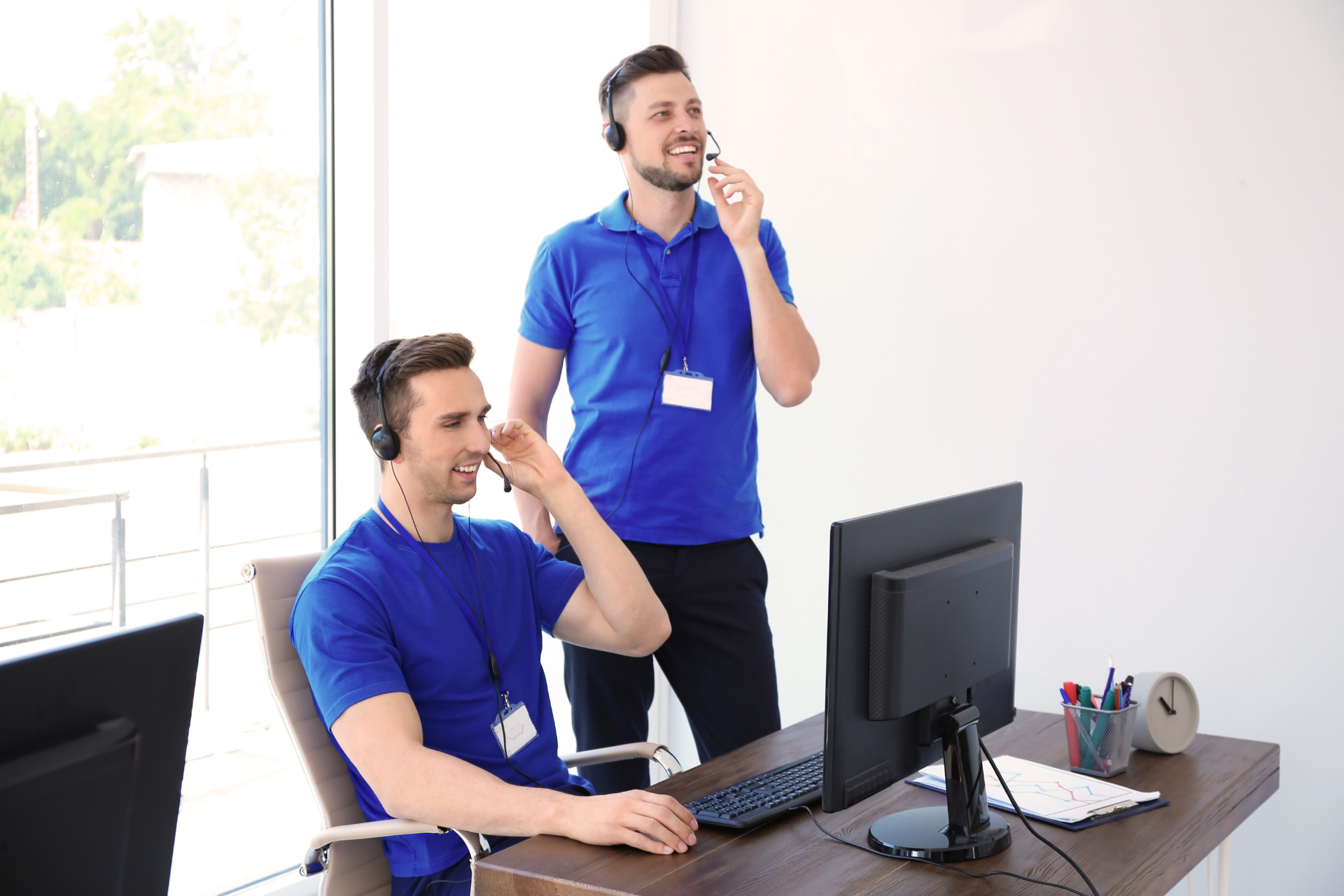 Two men in blue shirts with headsets in an office; one seated at a desk, the other standing, both smiling.