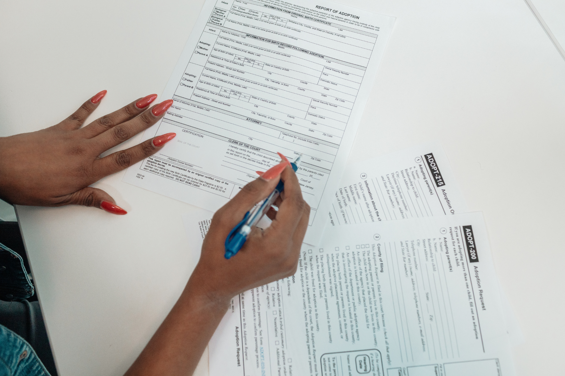 Hands with red nails filling out forms on a white surface with a blue pen.