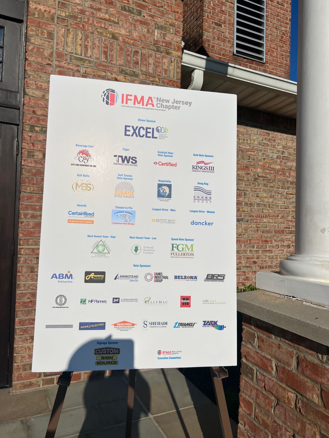 A white sign with logos on it is sitting on a wooden easel in front of a brick building.