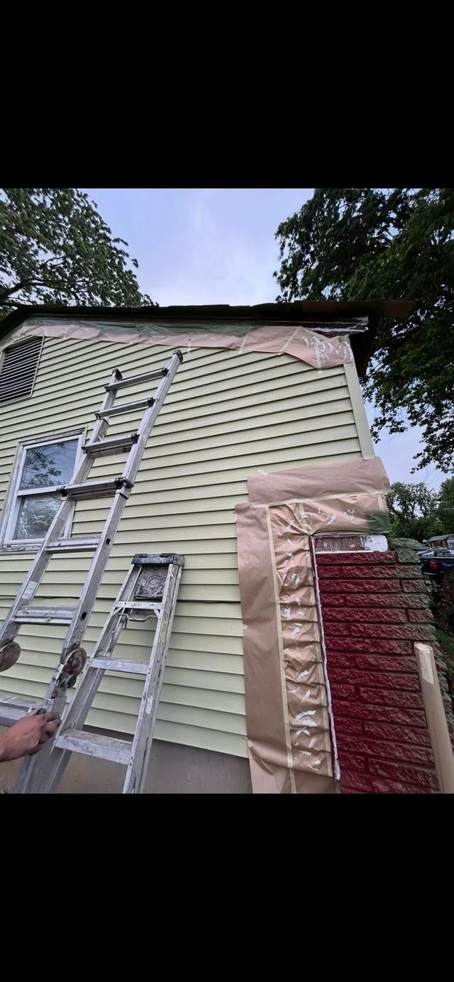 Ladders leaning against a beige house with siding, under trees and beside a brick chimney.
