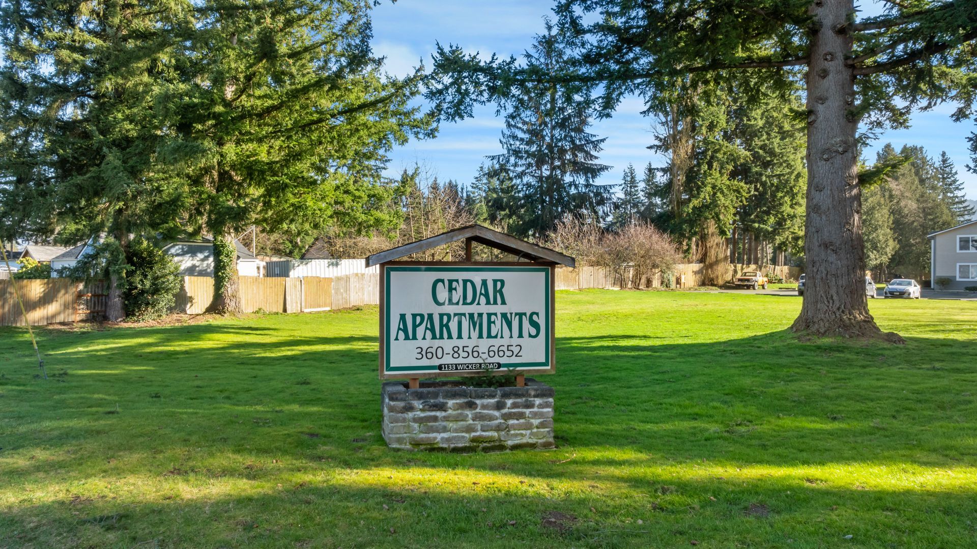 A sign for cedar apartments is in the middle of a grassy field.