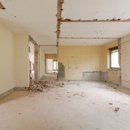 Interior of a room undergoing demolition with exposed brick, debris on the floor, and a window.