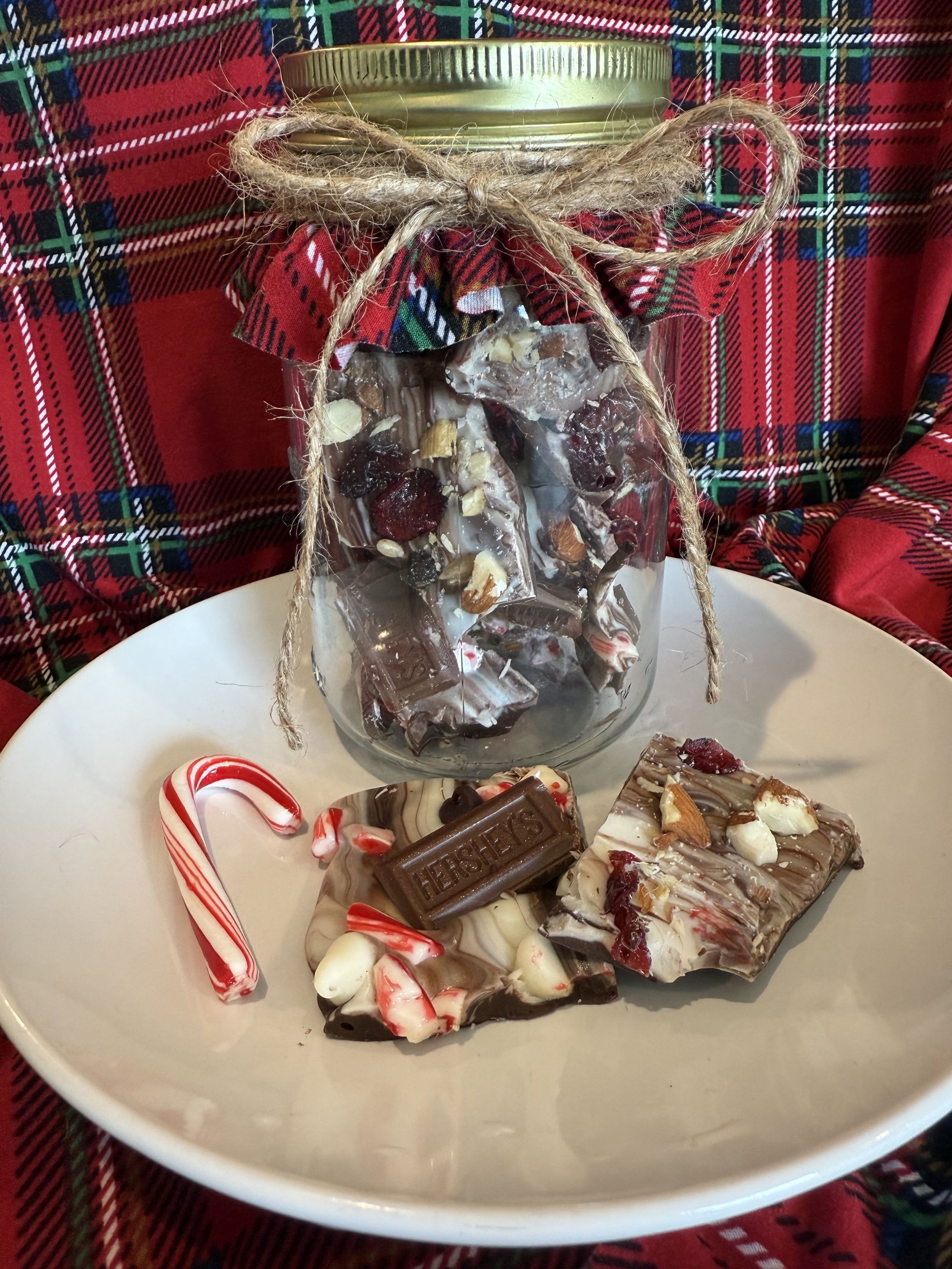 Jar of chocolate bark with candy cane on a white plate, set against plaid fabric.