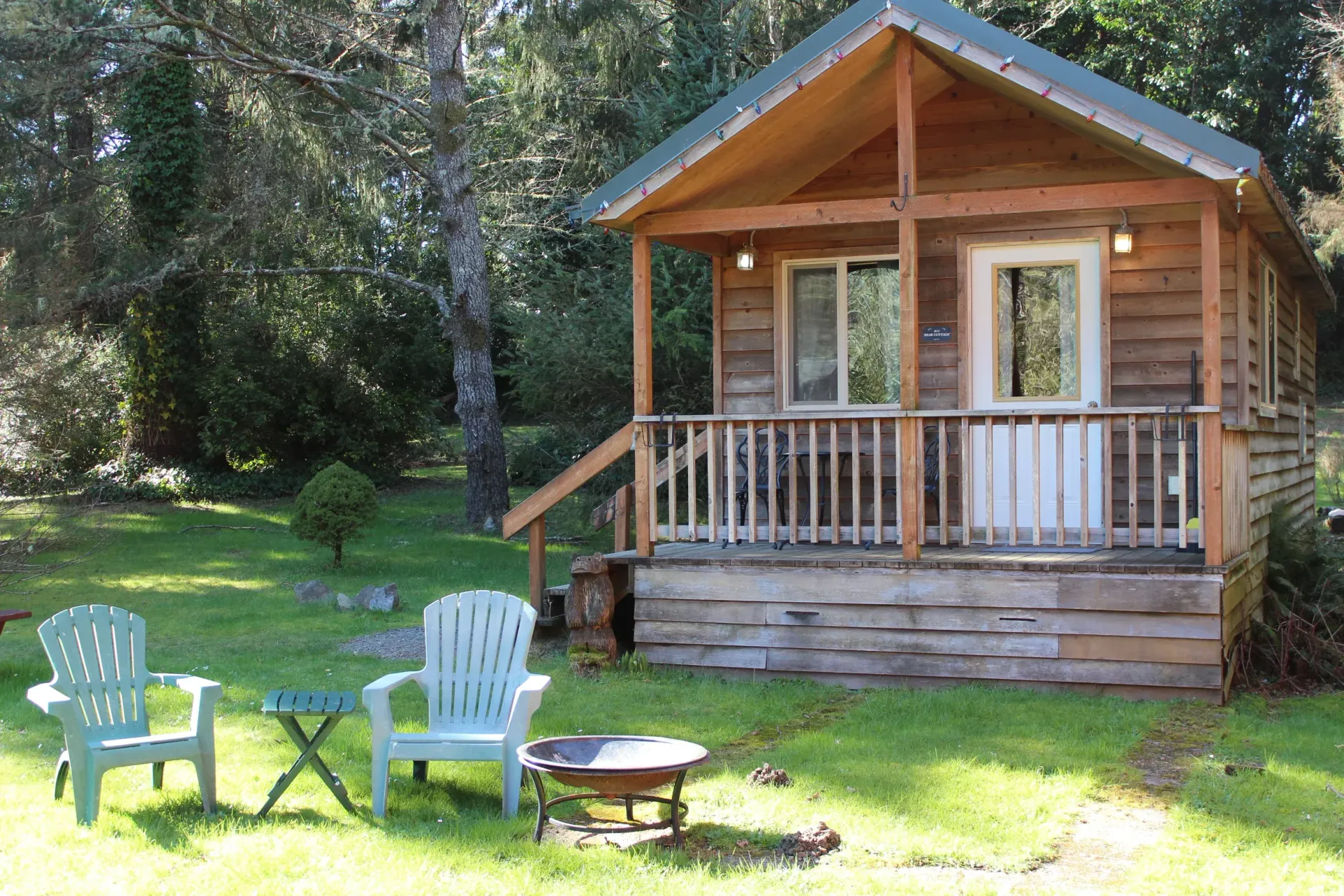 Wooden cabin with porch in a sunny wooded yard, with two chairs and a fire pit in front