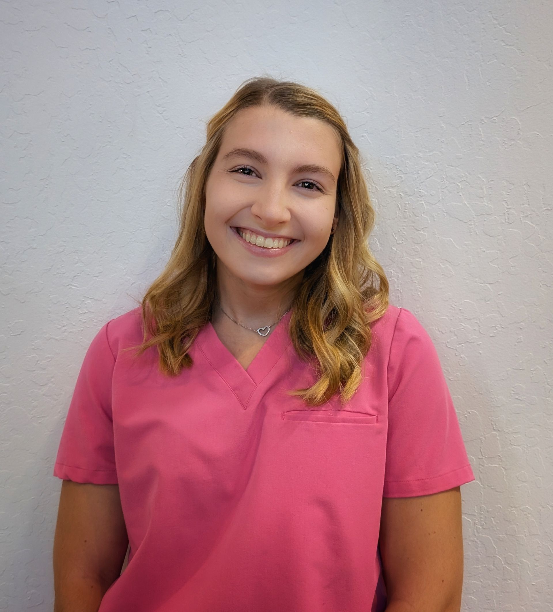 Smiling person in pink scrubs against a textured, light wall.