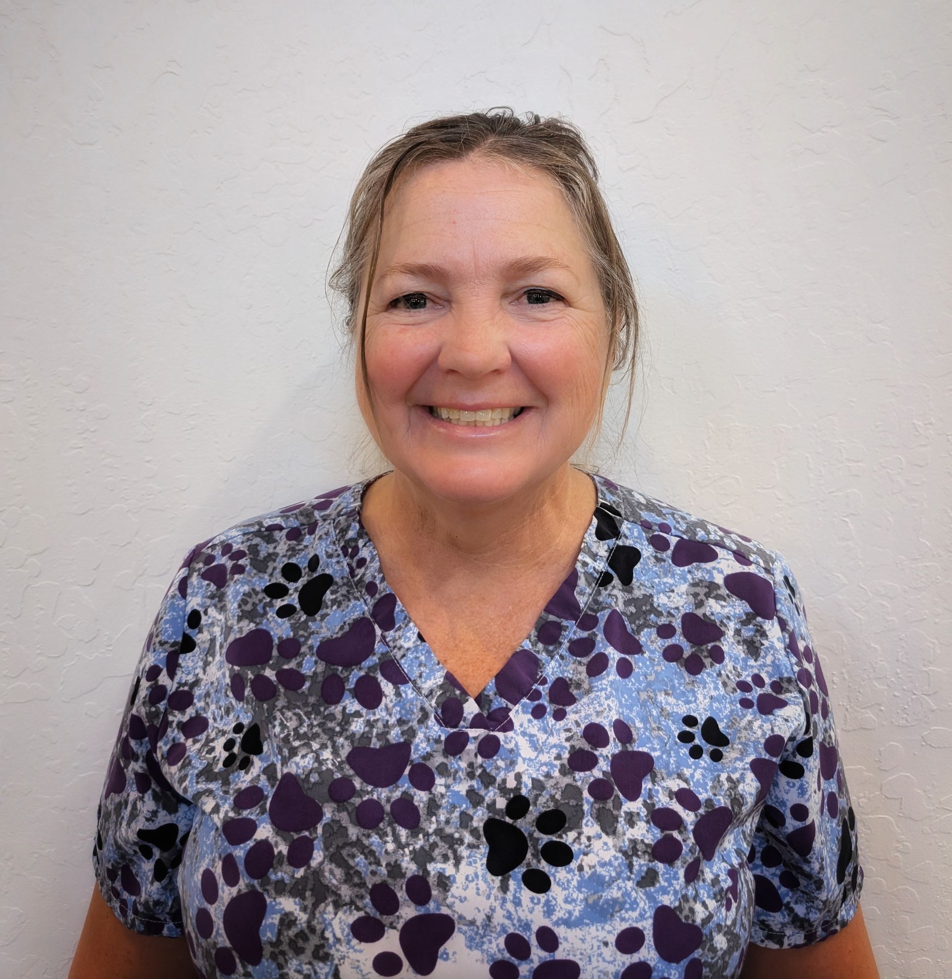 Woman in patterned scrubs smiles at the camera, standing against a white wall.