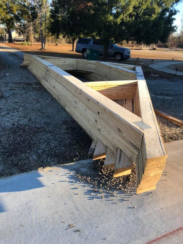 Wooden roof trusses stacked on a concrete surface. A truck is parked in the background.