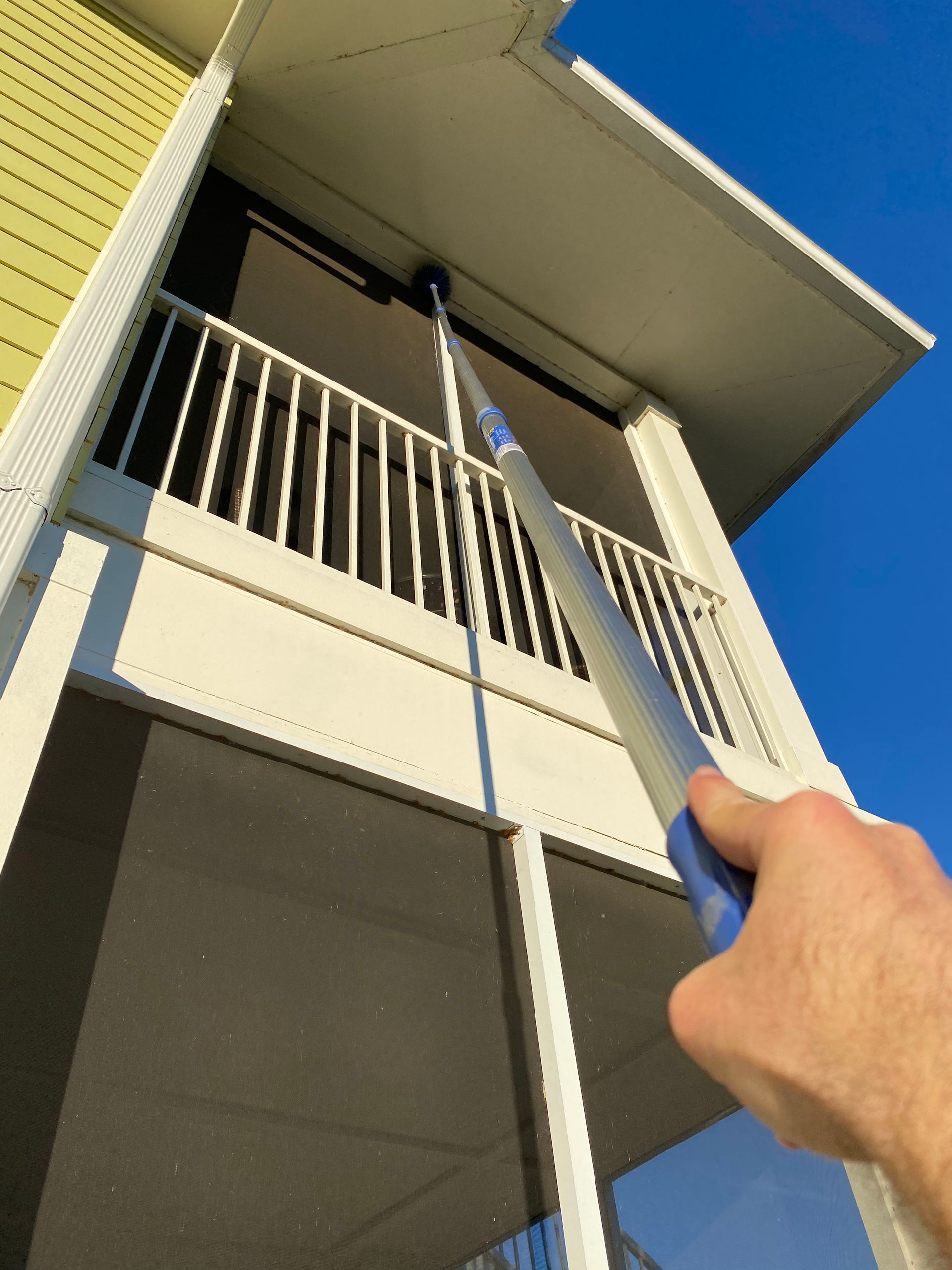 A person cleans a window screen with a long-handled tool on a balcony. Blue sky.