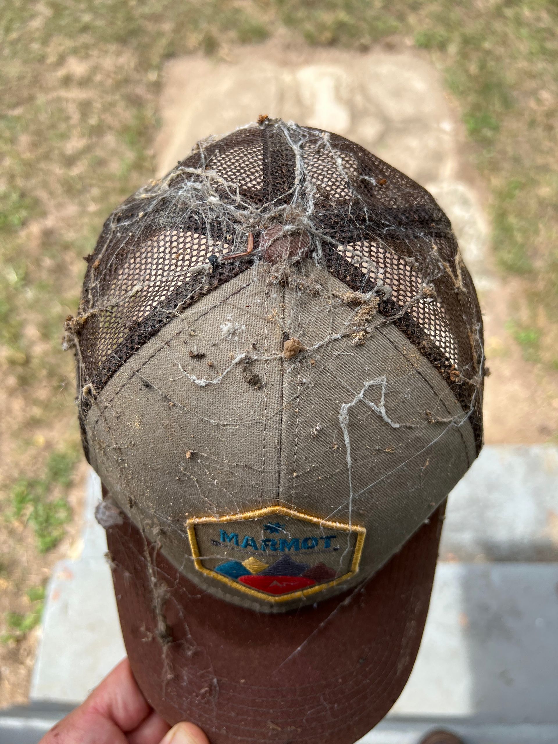 A dirty, worn Marmot baseball cap covered in cobwebs, held by a person outside.