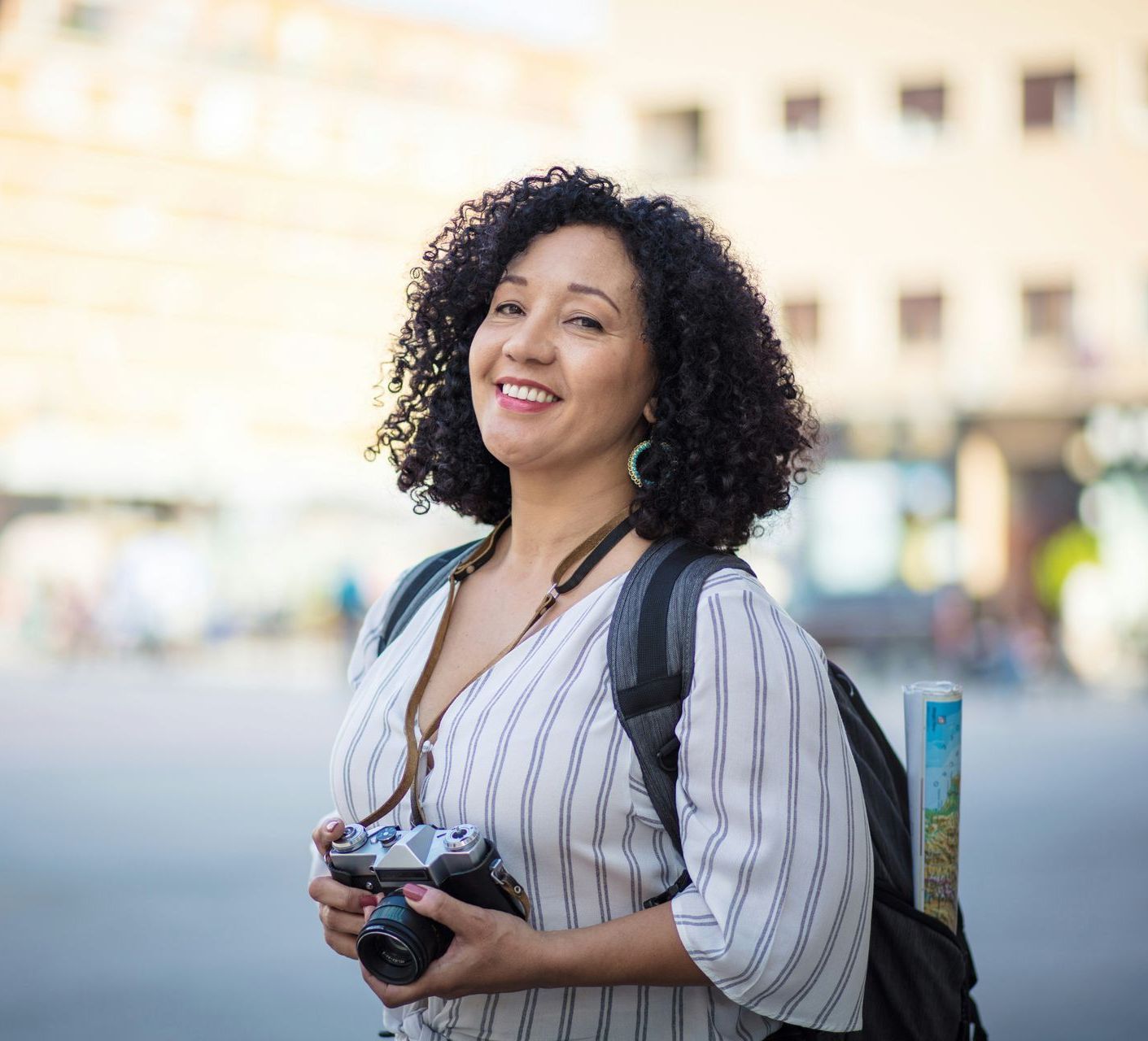 Woman with curly hair smiling, holding a camera and wearing a backpack, outdoors.