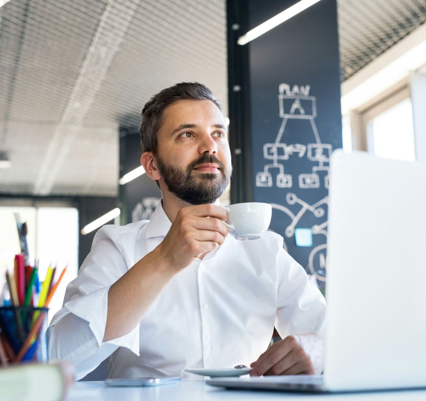 Man with a beard sips from a coffee cup while looking up, sitting at a desk with a laptop, in an office setting.
