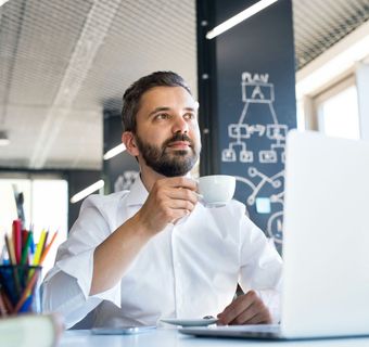 Man with a beard sips from a coffee cup while looking up, sitting at a desk with a laptop, in an office setting.