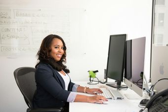 Woman in a blazer smiles while working at a desk with dual monitors, a keyboard, and a phone in a bright office.