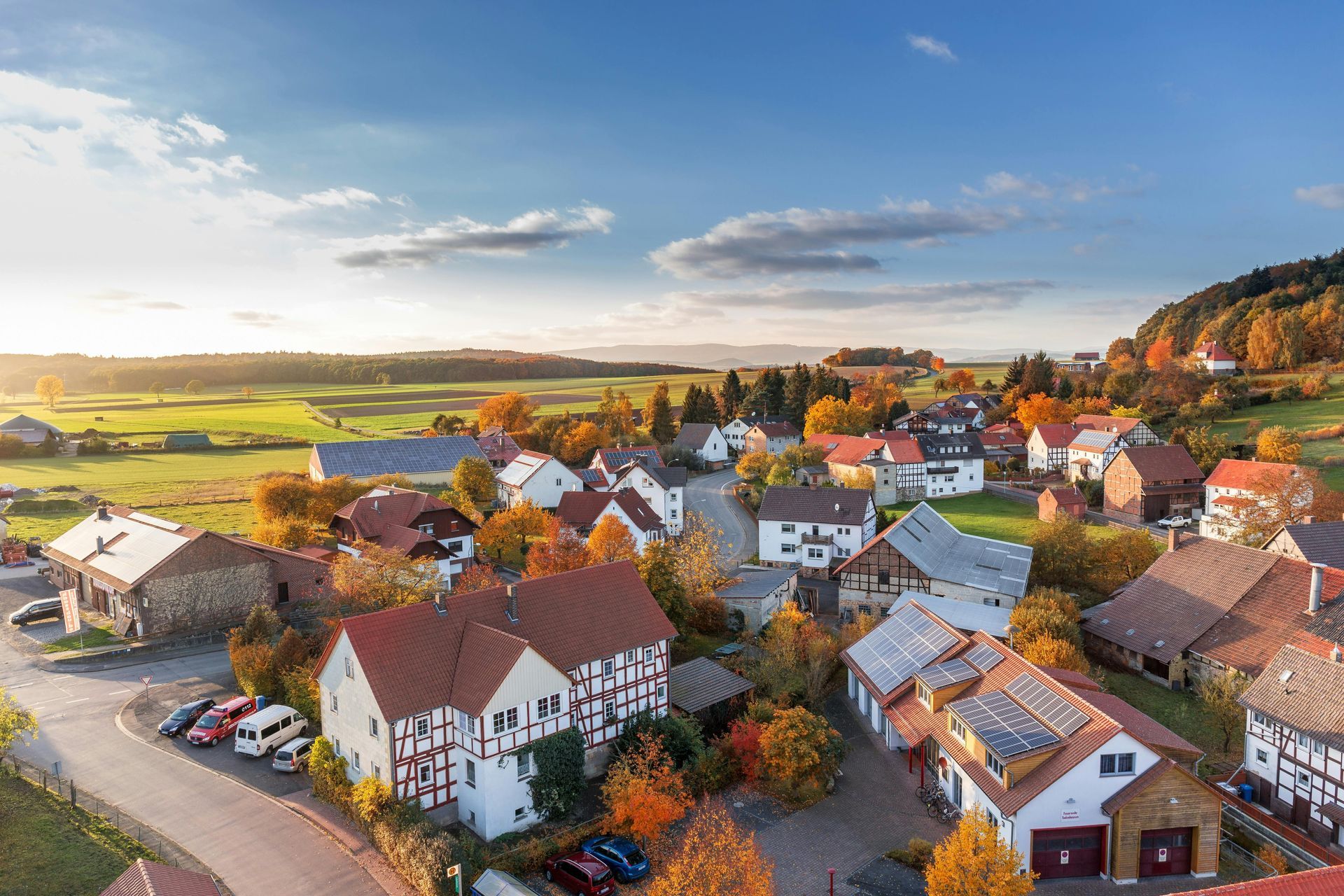 Aerial view of a small village with red-tiled roofs, surrounded by autumn foliage and fields, under a bright blue sky.