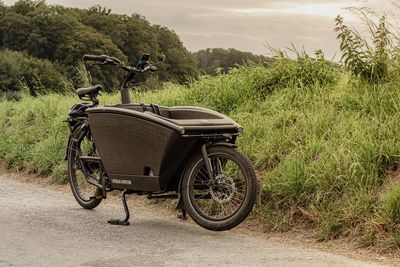 Een zwarte bakfiets staat geparkeerd op een grindpad naast een grasheuvel.