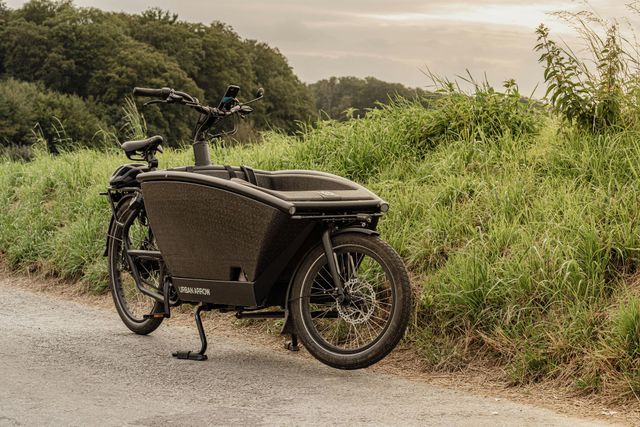 Een zwarte bakfiets staat geparkeerd op een grindpad naast een grasheuvel.