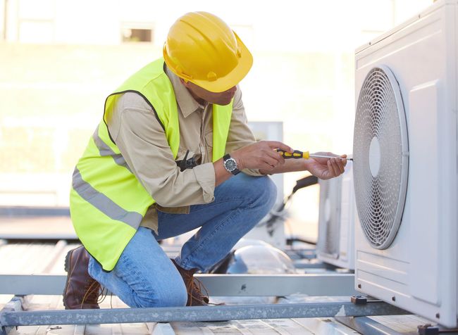 Technician in yellow vest and hard hat inspecting an outdoor HVAC unit with a tool