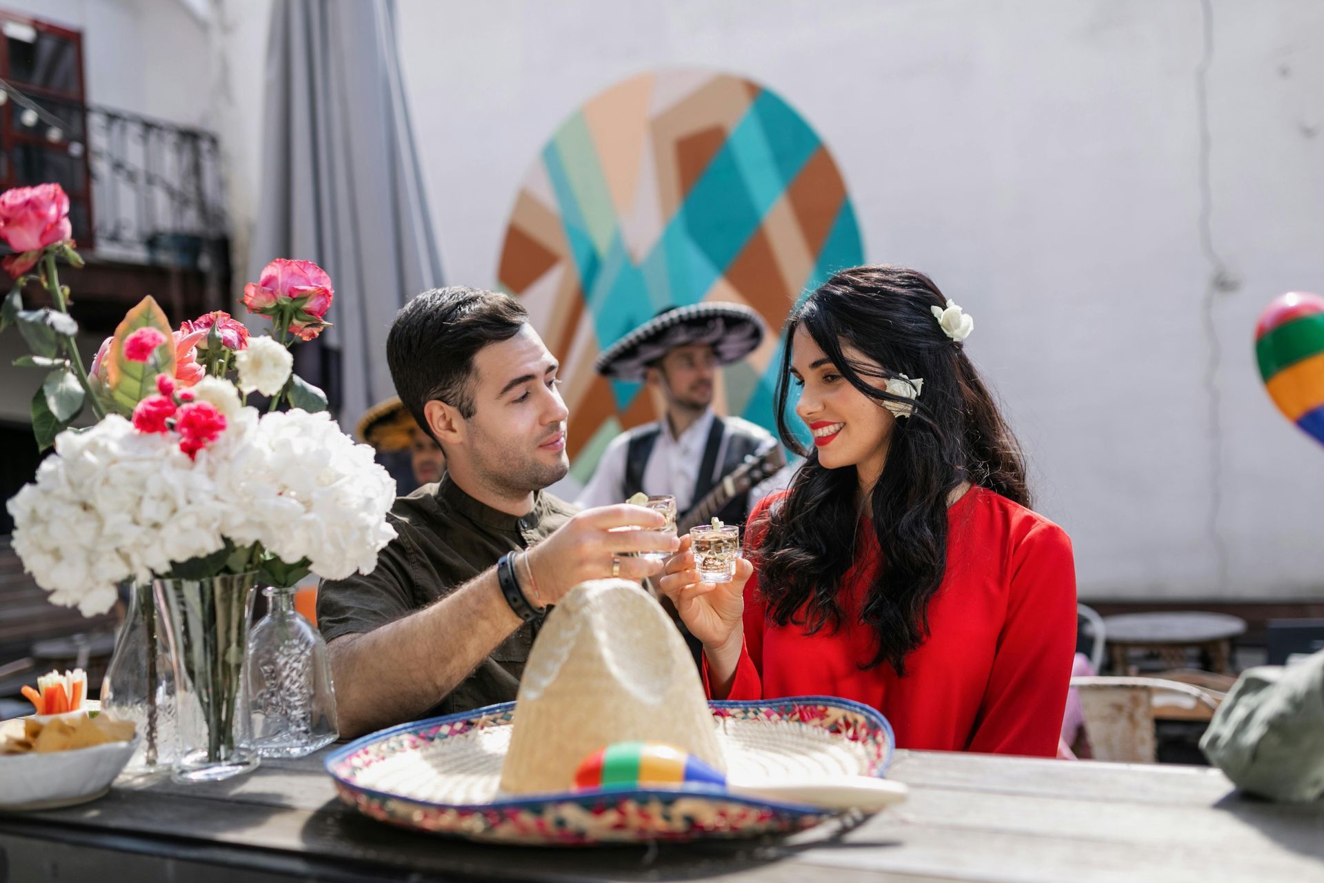 Two people smile and clink drinks at a table with flowers and a sombrero in a festive setting