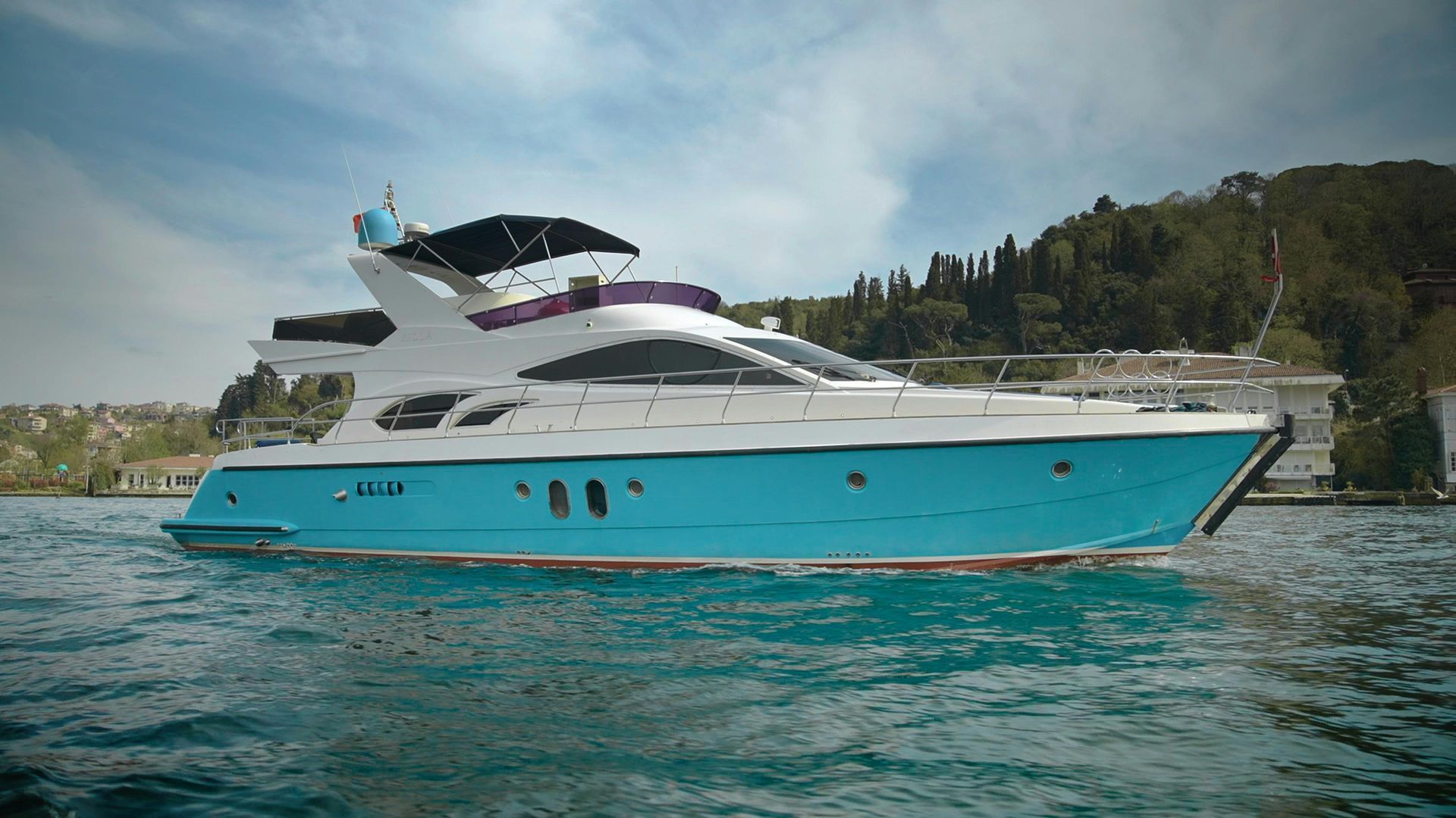 Turquoise and white yacht cruising on calm water near a wooded shoreline.