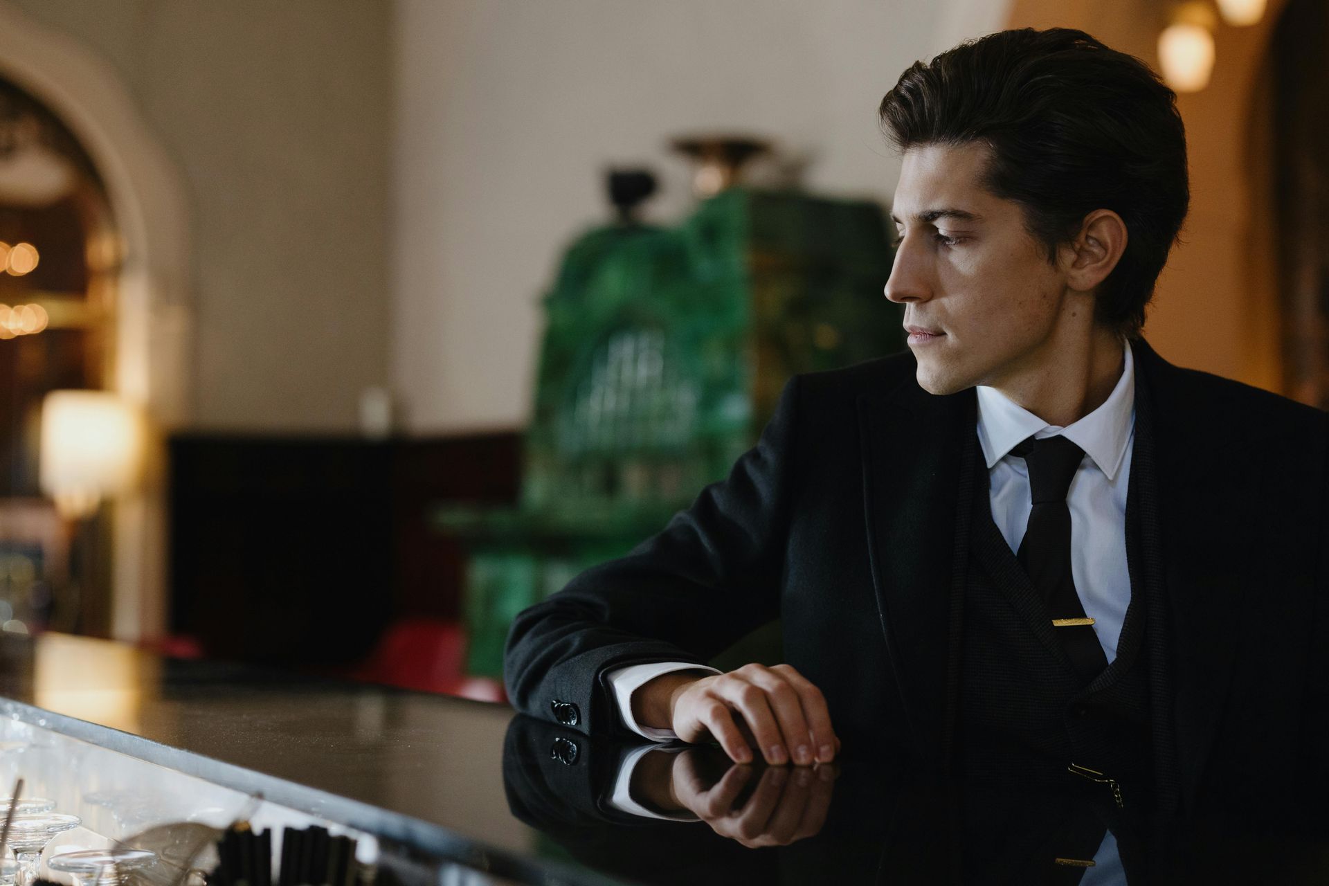Man in a dark suit sitting at a bar, looking to the right in a warmly lit room.