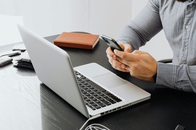Person using a smartphone beside an open laptop on a desk with a notebook.