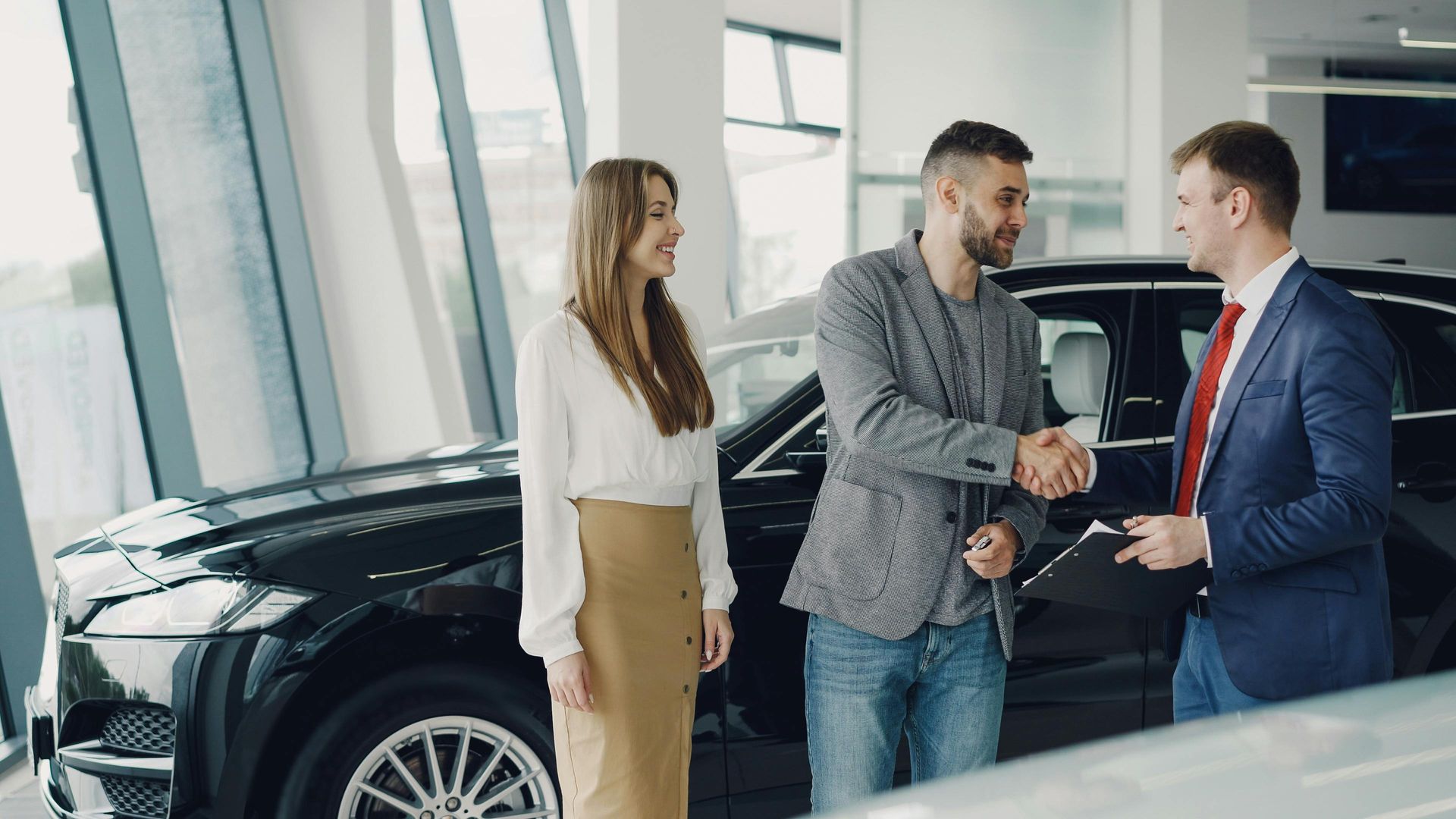 Car dealership handshake beside a black car, with a woman standing nearby in a bright showroom