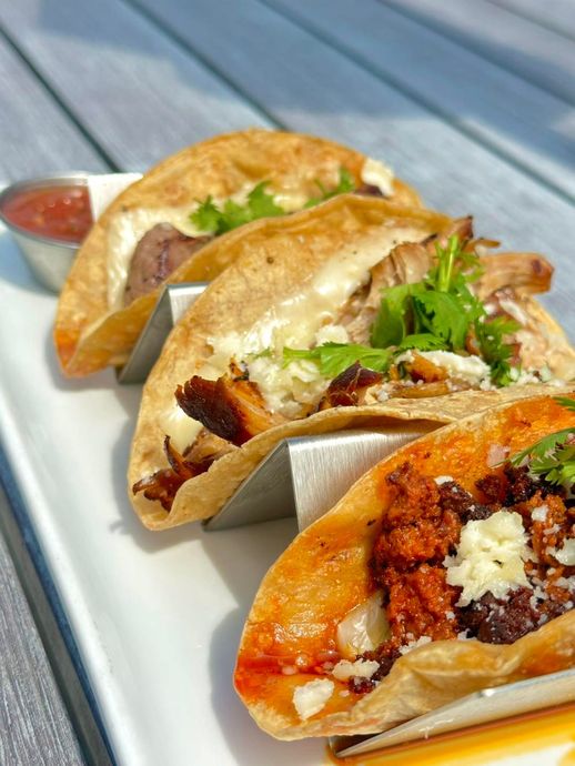 Four tacos on a white plate with metal holders, topped with meat, cilantro, and cheese, with a small salsa bowl.
