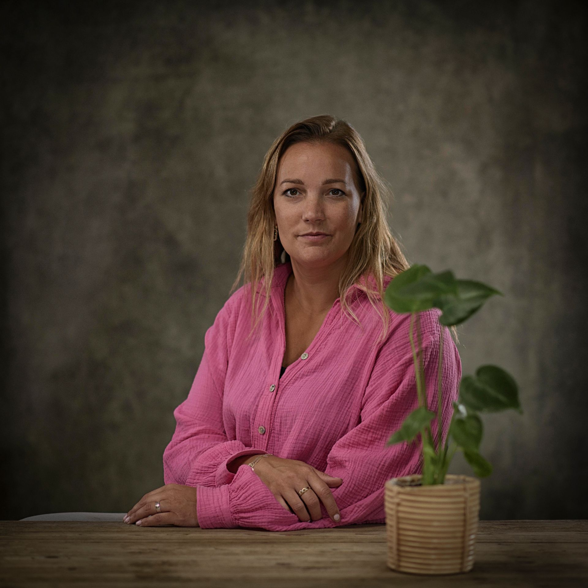 Een vrouw in een roze shirt zit aan een tafel met een potplant.