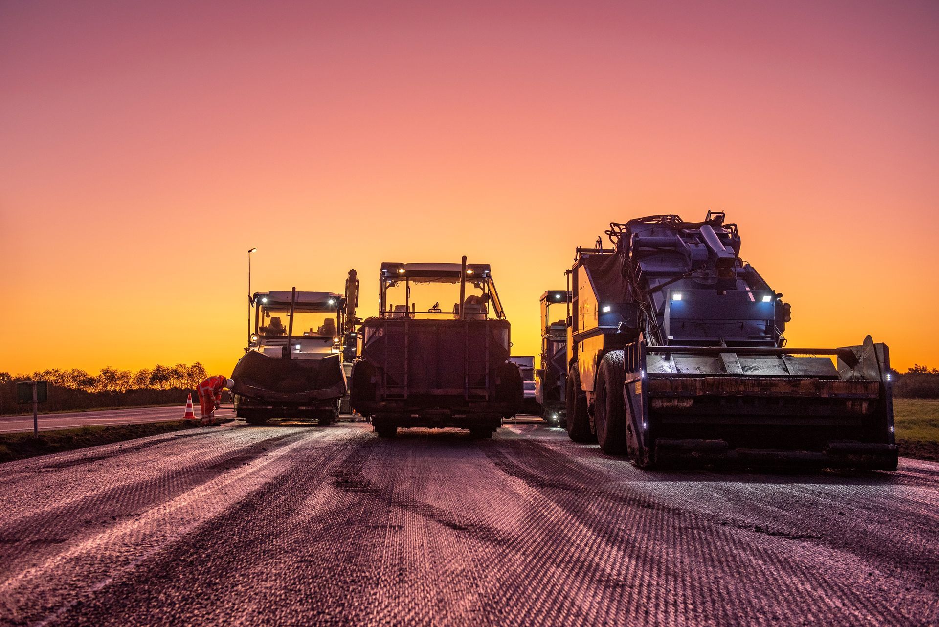 Een groep bouwvoertuigen rijdt bij zonsondergang over een weg.