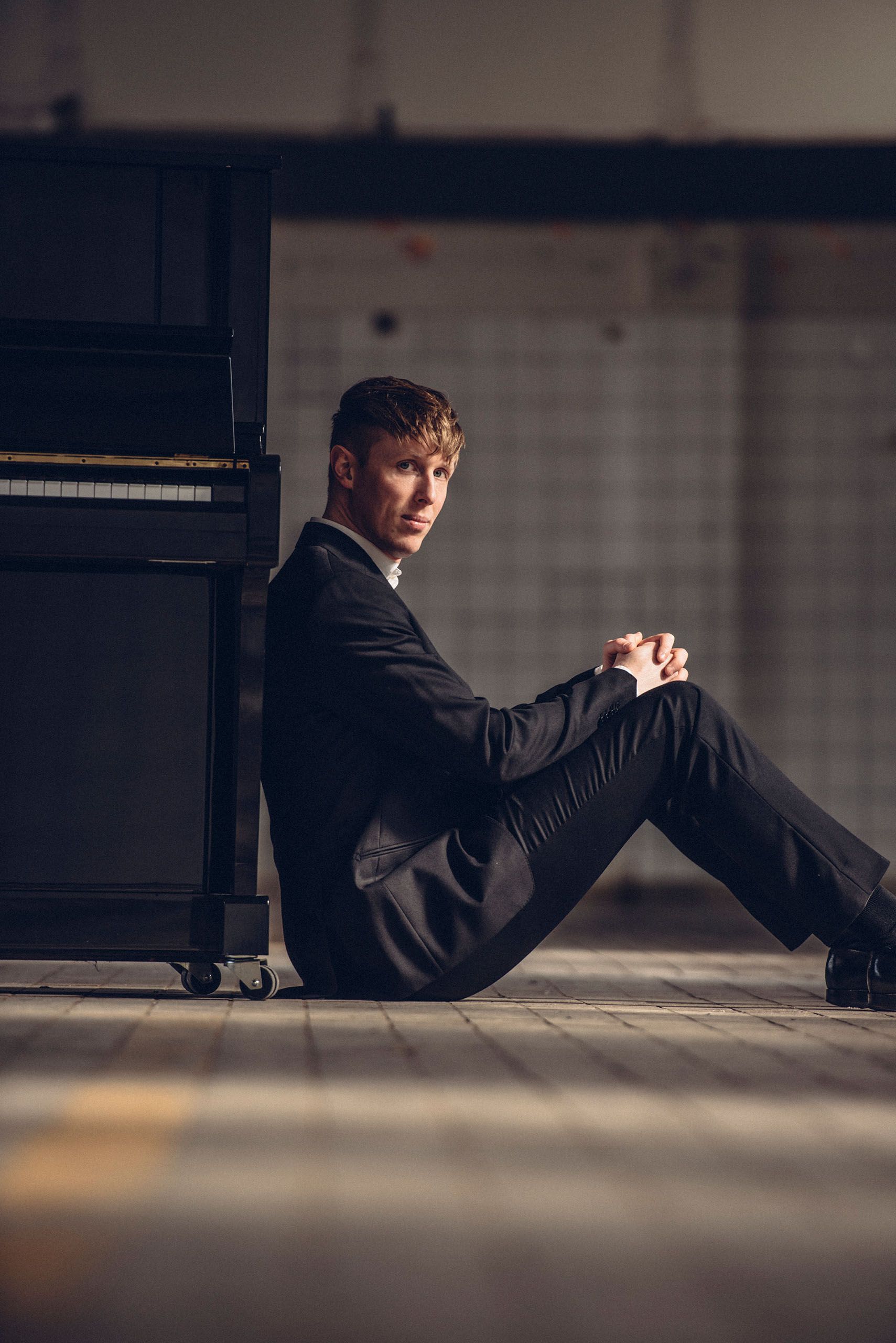 Een man in pak zit op de grond naast een piano.