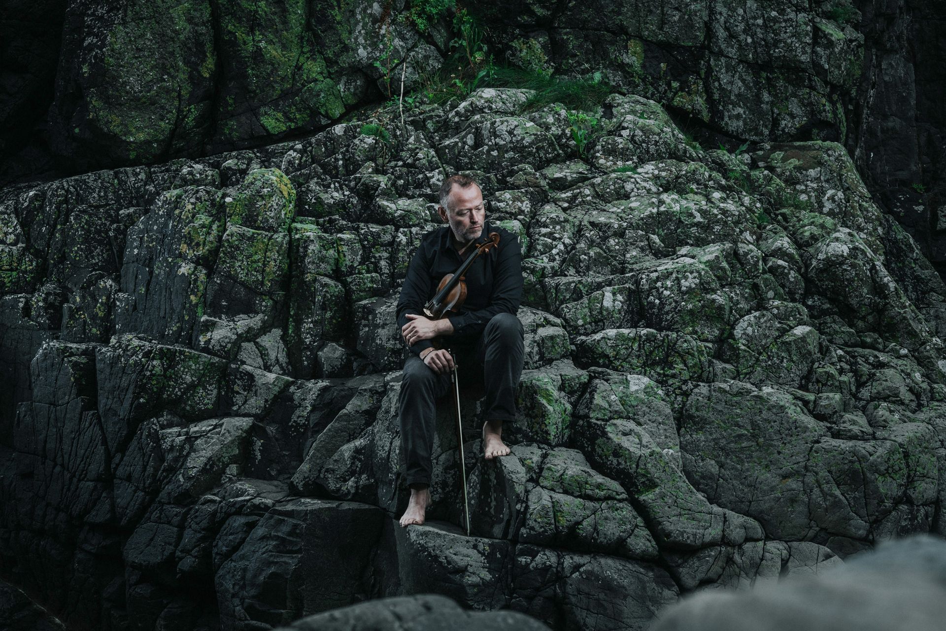 Een man zit op een rots en houdt een gitaar vast.