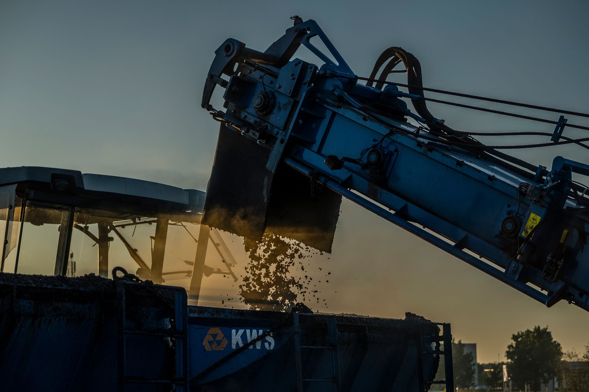 Een grote blauwe kraan laadt een afvalcontainer bij zonsondergang.