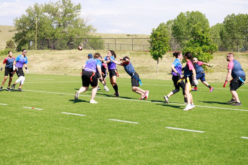 Flag Football players in blue jerseys compete on a grassy field near trees and a fence.