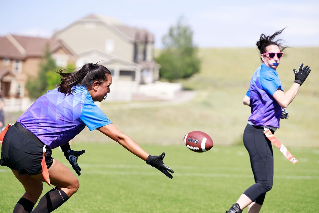 Two athletes in blue jerseys reaching for a football on a grassy field with houses in the background