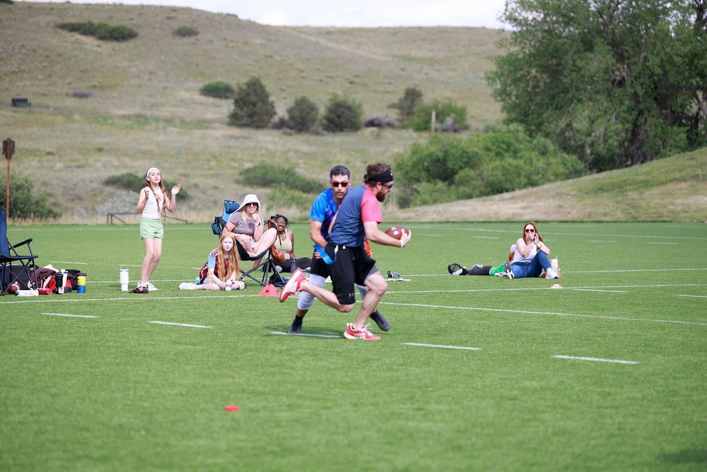 Players sprint across a grassy field during a sports game, with spectators and hills in the background.
