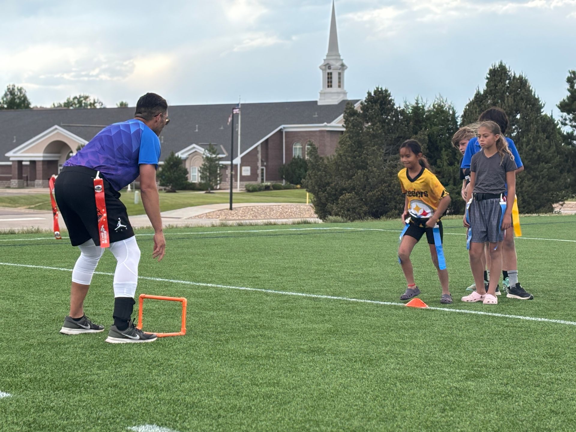 Coach setting up a drill for three kids on a grassy field with an orange hoop and church buildings in the background