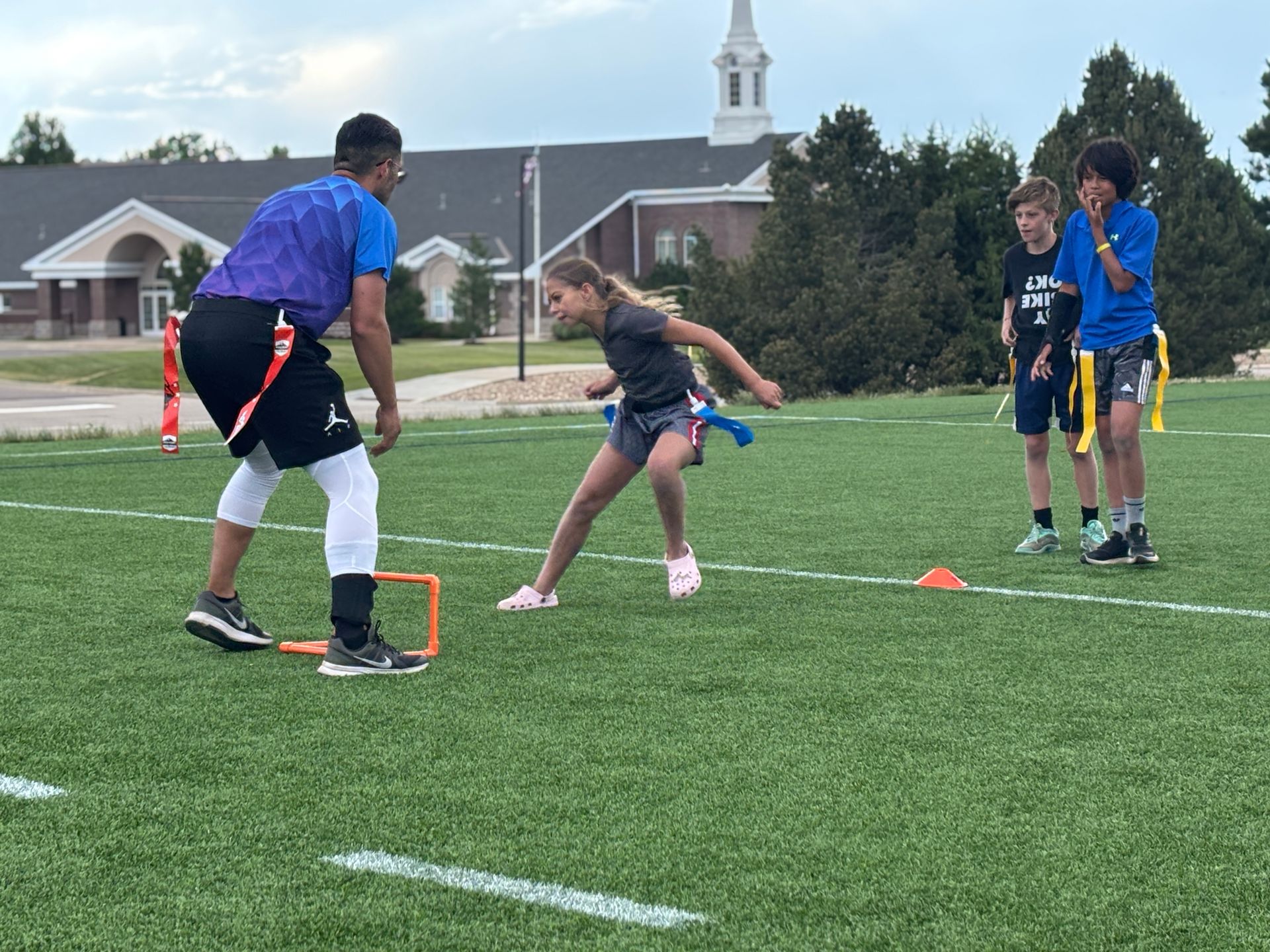 Youth flag football drill on a turf field, with a girl dodging a coach and teammates watching nearby.