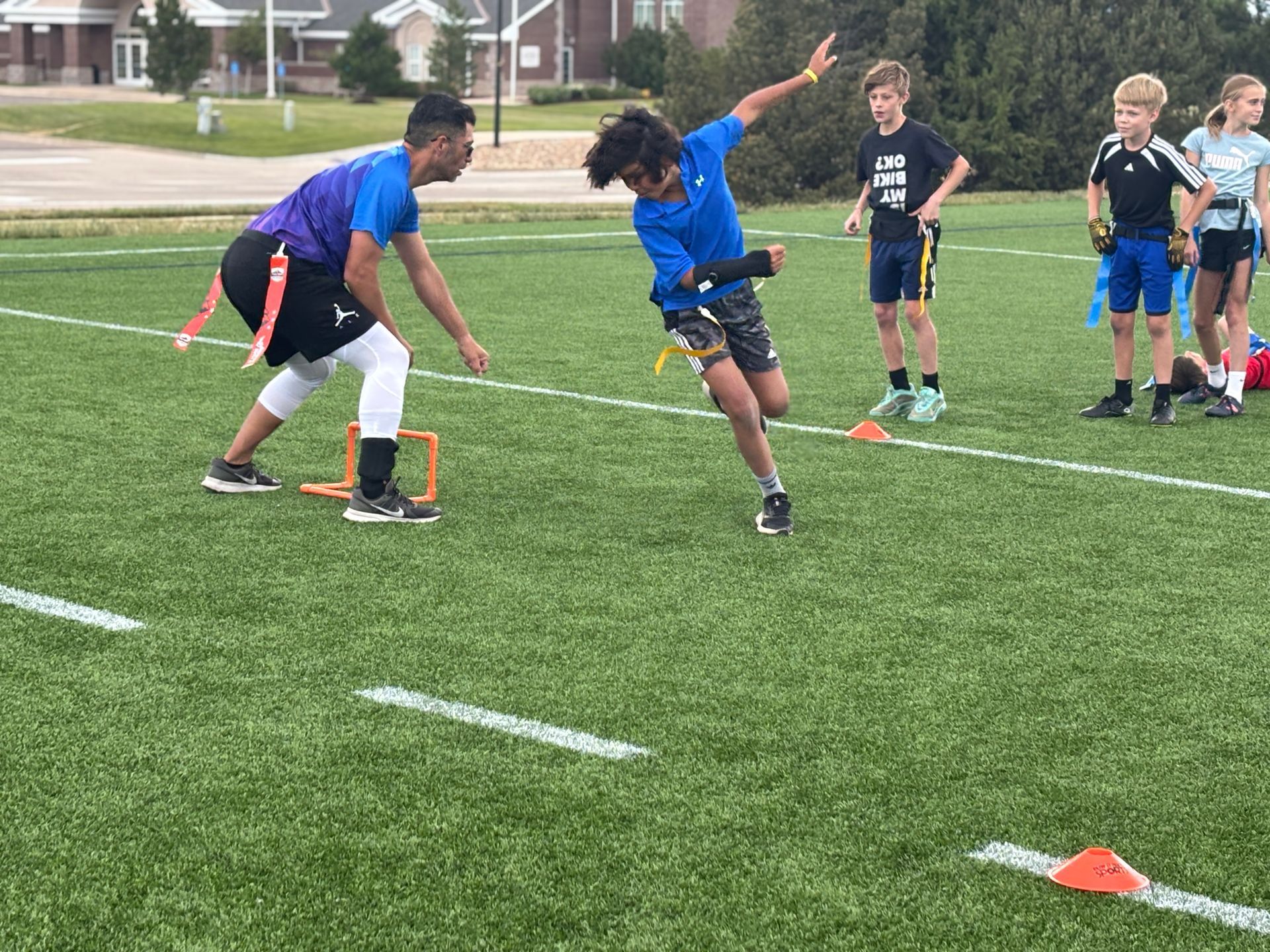 Children doing a relay race drill on a grassy field with orange cones and a coach supervising