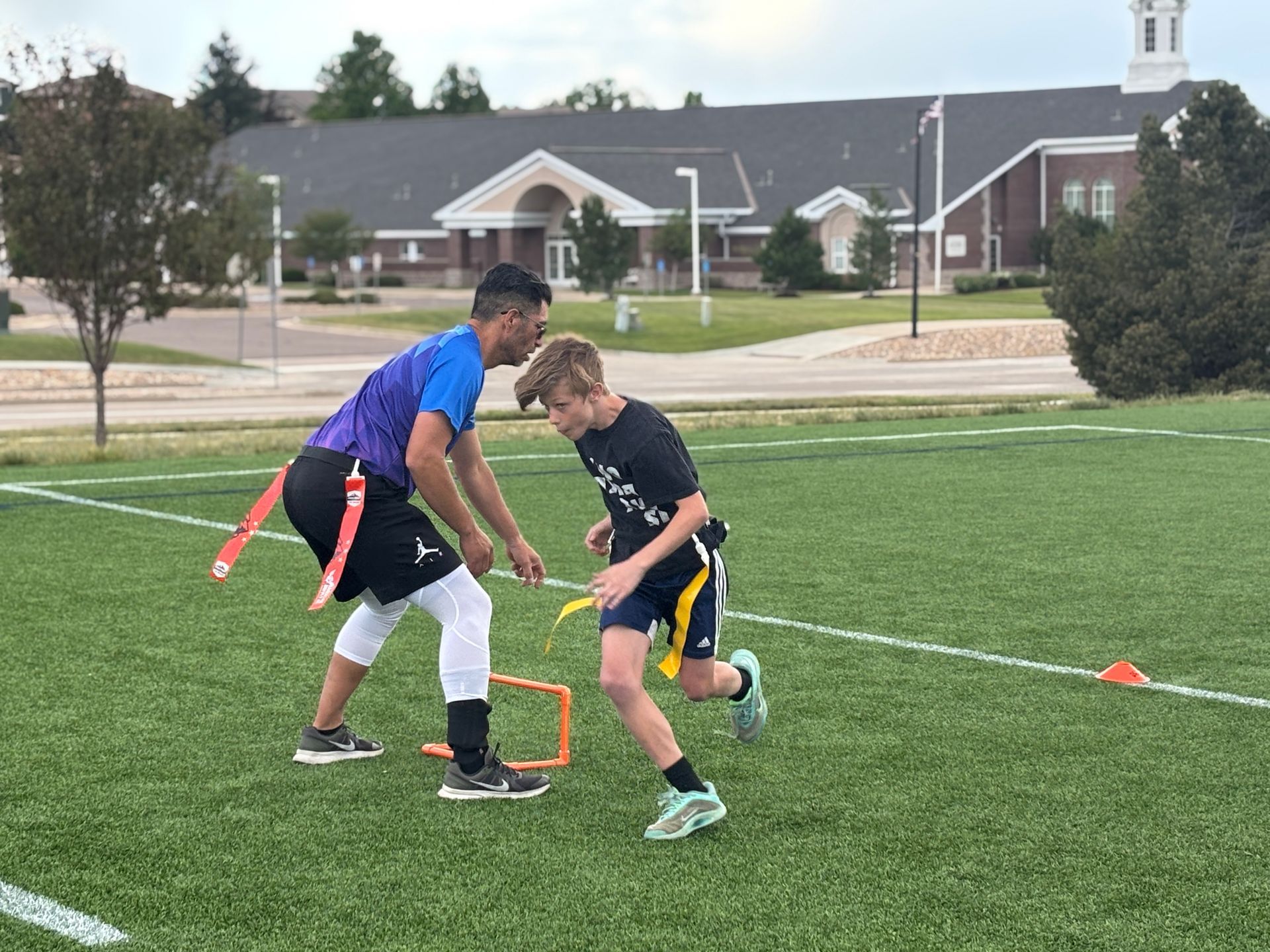 Two children playing flag football on a green field with a small orange hurdle