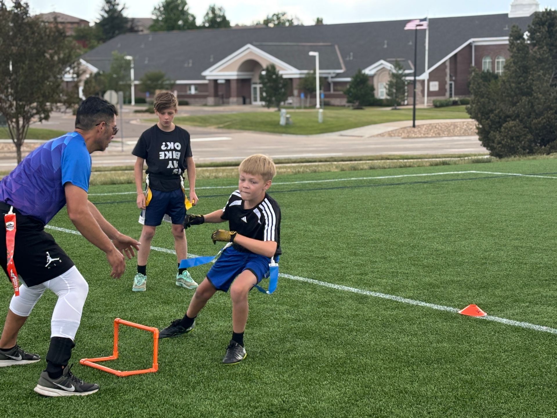 Youth soccer drill on a grassy field, coach guiding a boy kicking a ball near orange training hurdles.
