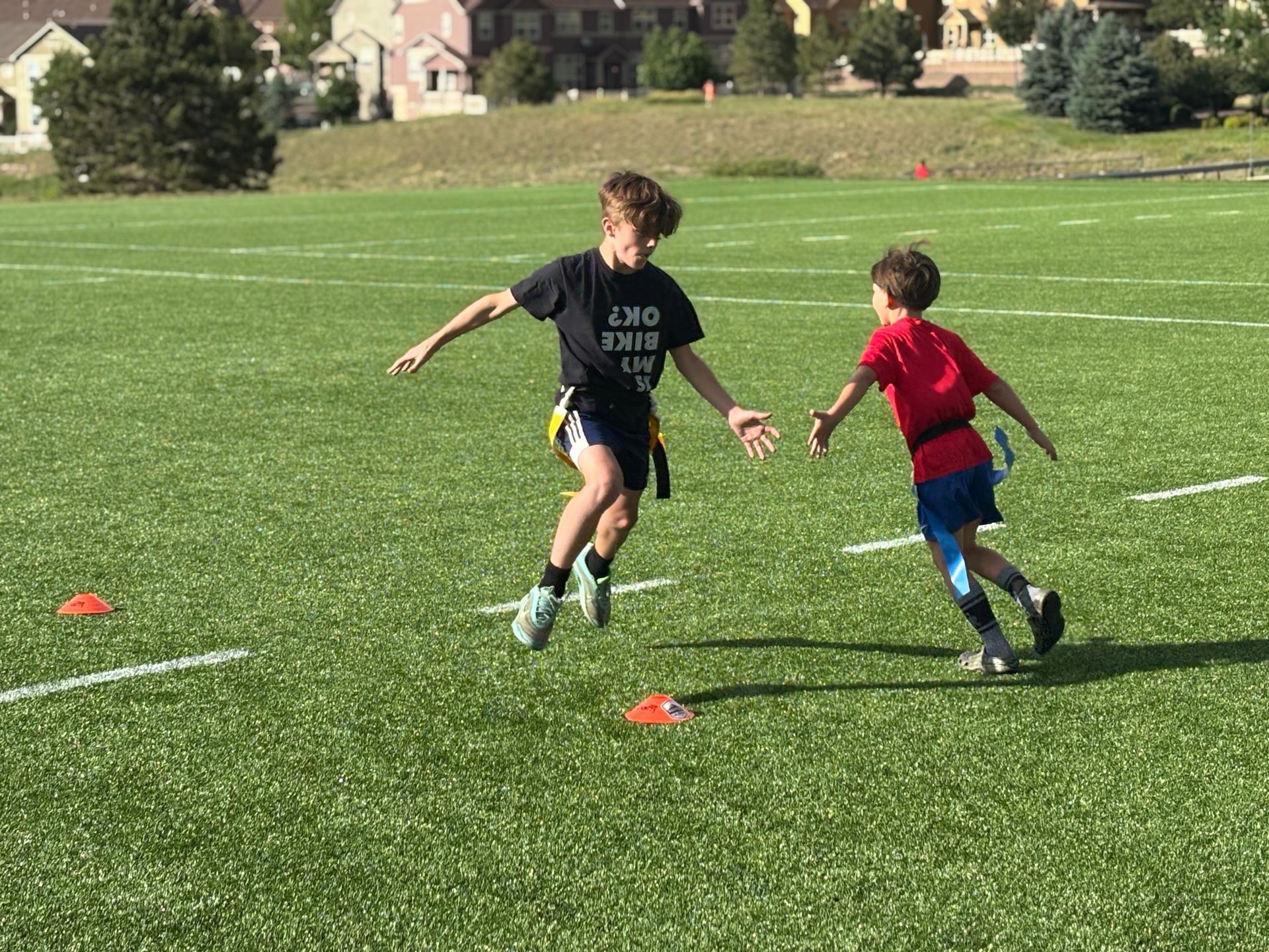 Two children in black and red shirts running on a grassy field between orange cones.