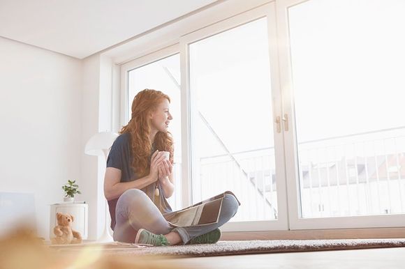 A woman is sitting on the floor in front of a window with a cup of coffee and a laptop.