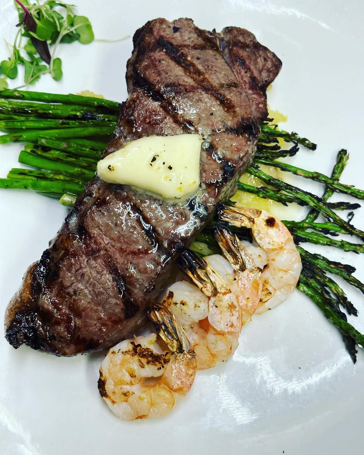 Grilled steak topped with butter, asparagus, and shrimp on a white plate.