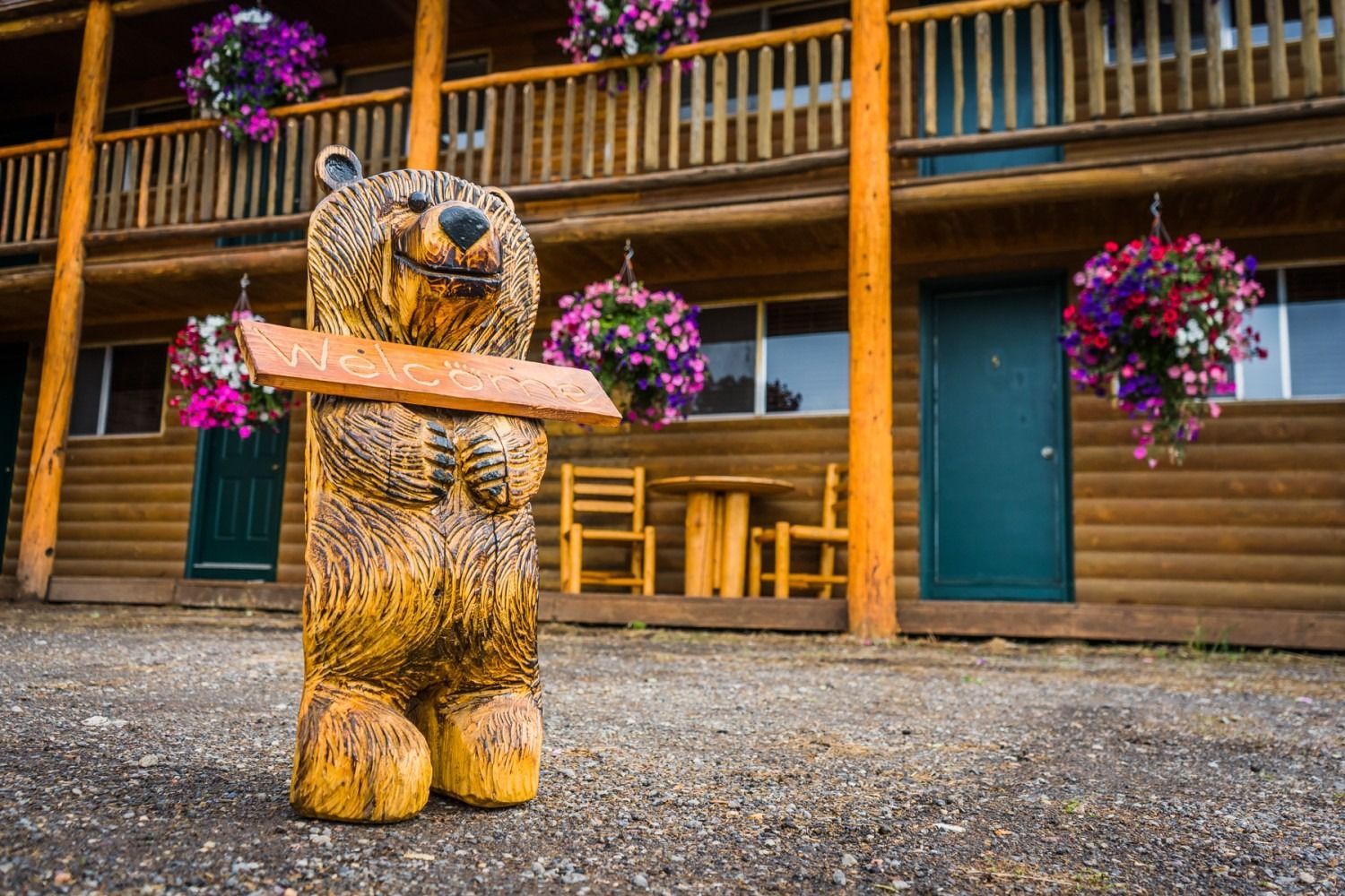 Carved wooden bear statue holding a sign outside a rustic lodge decorated with hanging flower baskets.