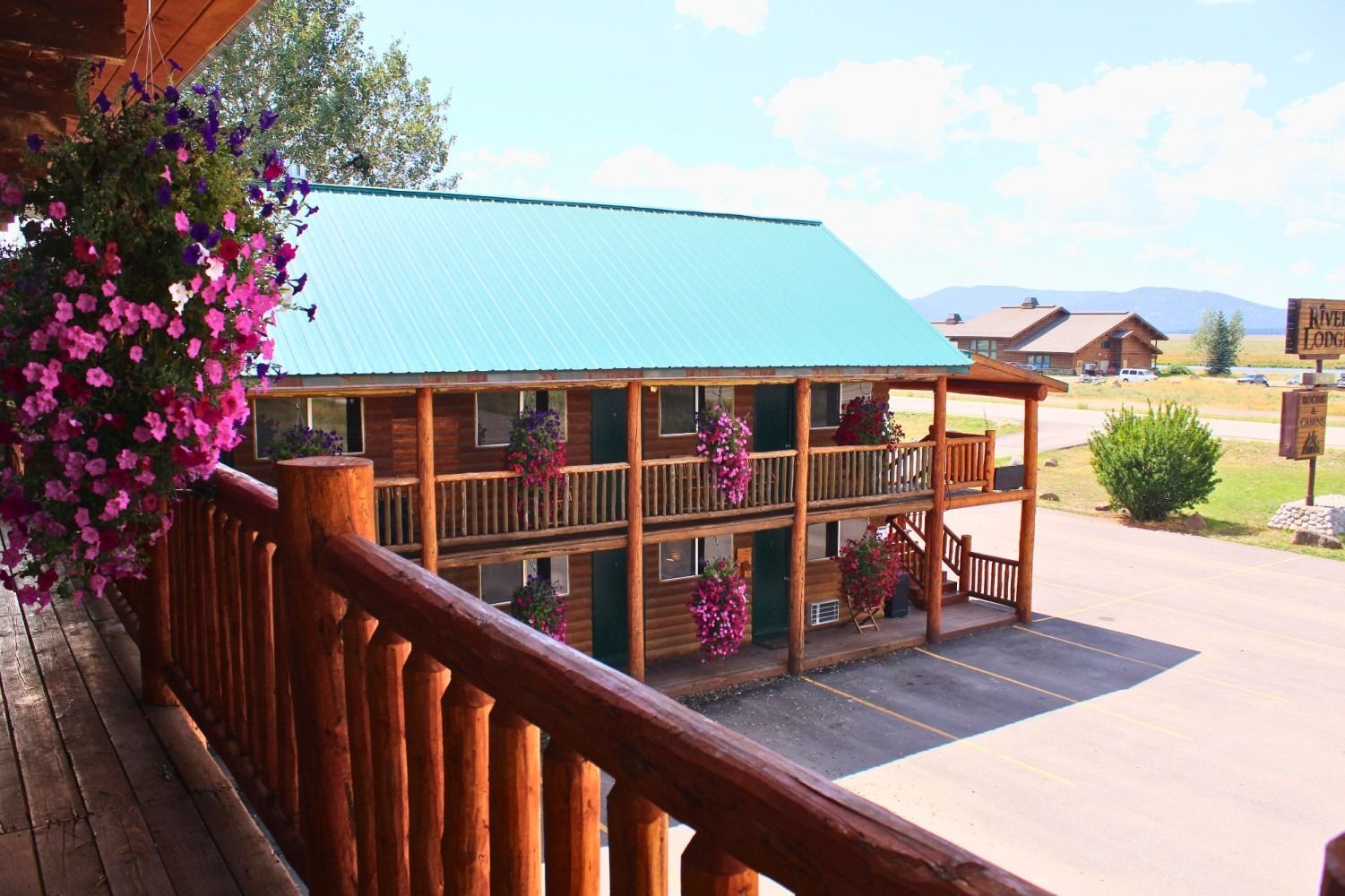 Two-story log motel with teal roof, porch with hanging pink flowers, and mountains in the background.