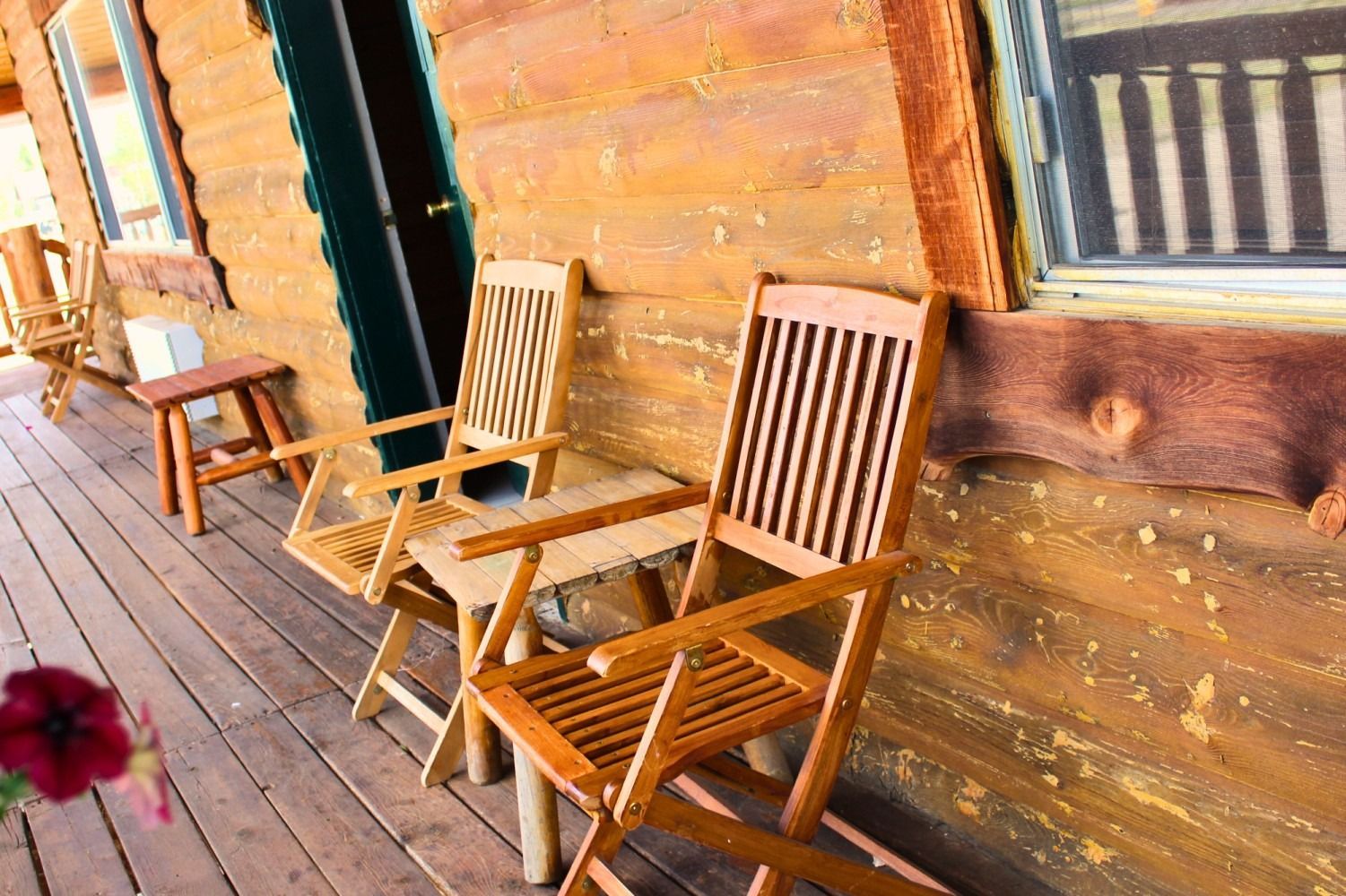 Wooden chairs on a porch of a cabin with a window and a small table.