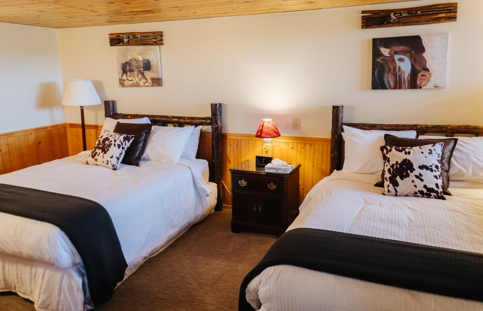 Bedroom with two beds, wooden headboards, cowhide pillows, artwork, bedside table, and lamps.