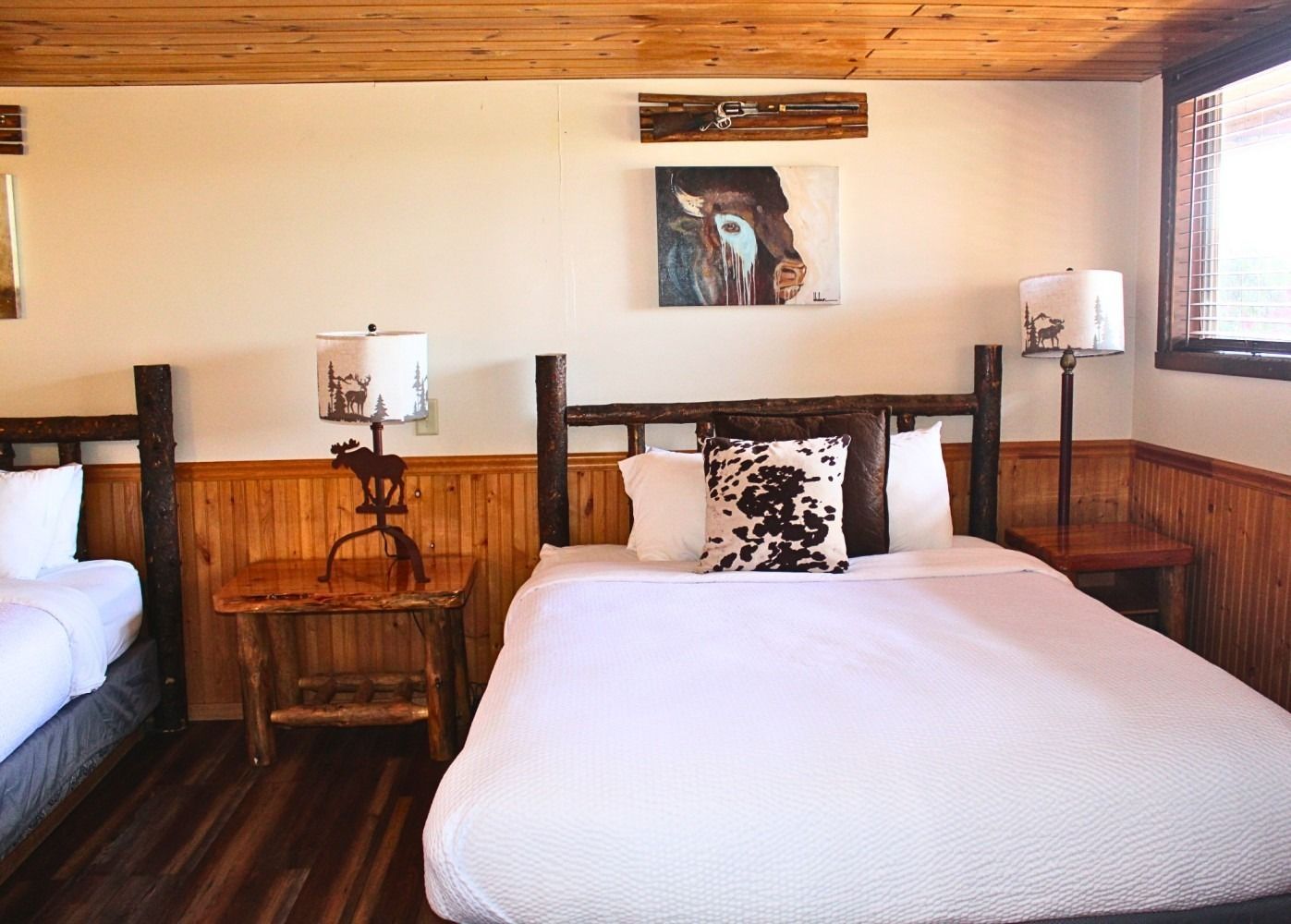 Bedroom with two beds, rustic wooden frames, wood paneled walls, white bedding, nightstands, and lamps.