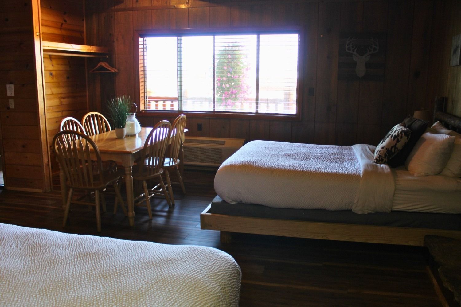 Cozy cabin interior with beds, dining table, and large window overlooking trees. Brown wood walls and floors.